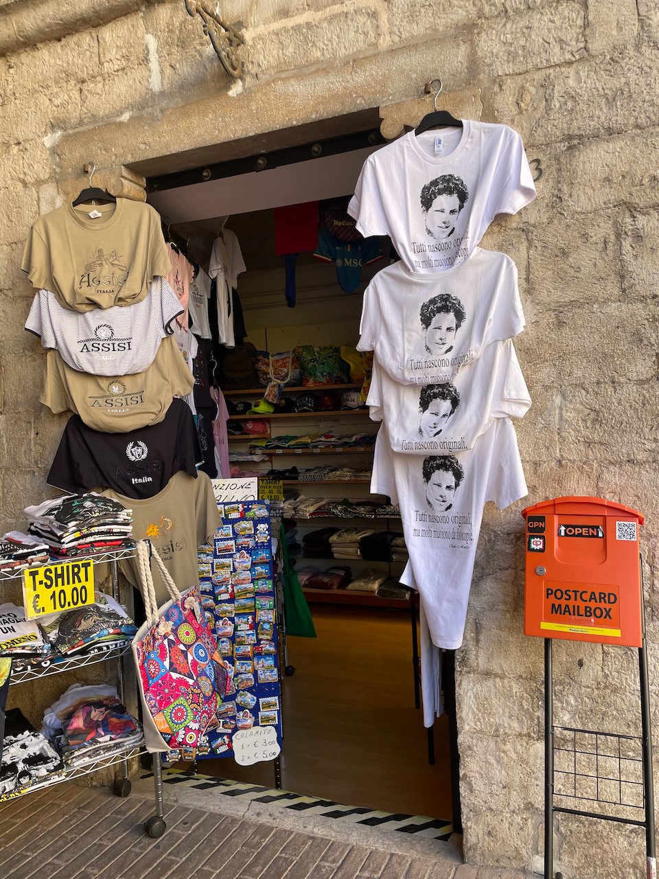 A souvenir stand in a doorway in Assisi with t-shirts with Carlo's picture and a quote