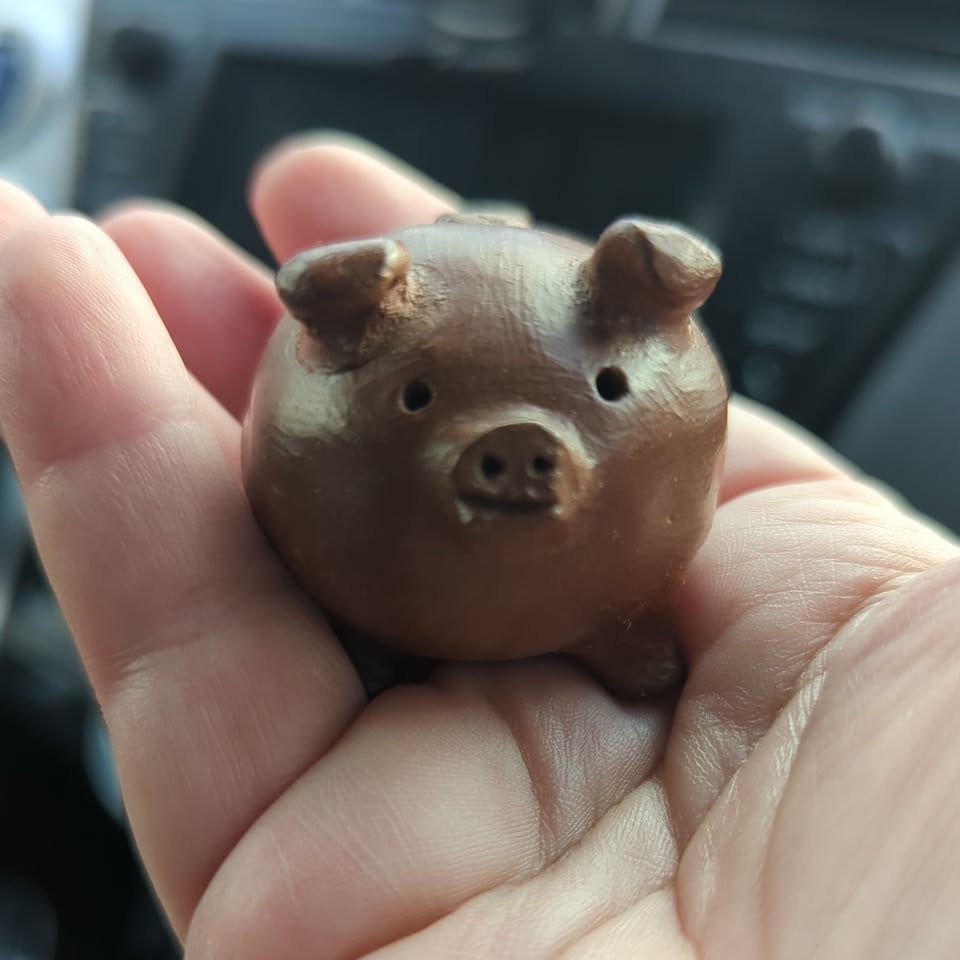A small ceramic pig rests in the palm of a hand. The pig is brown and has wide innocent eyes as it stares at the camera..