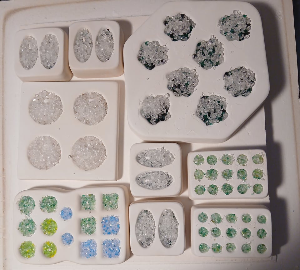 kiln shelf with a bunch of molds filled with glass frit, mostly circles and ovals and some paws