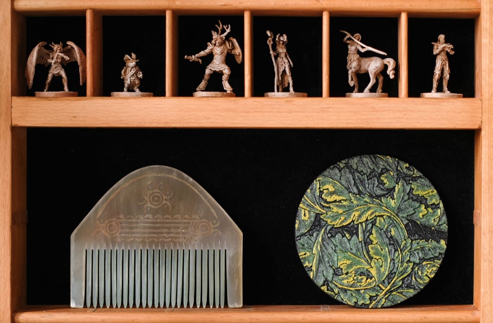 Photo close-up of the compartments and the trinkets inside. A variety of beige-painted fantasy RPG figurines line the small compartments on top, and one big compartment below contains an antler bone viking haircomb and a round 3D-printed coaster with a leafy motiv.