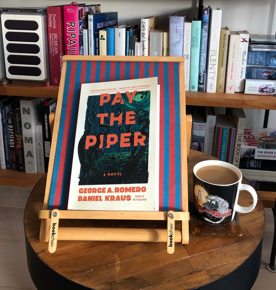 A book, Pay the Piper, on a red and blue striped book chair next to a mug of coffee with a scary gremlin on it. Bookcase in the background.