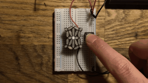 An animated gif showing a small custom-built LED display. It has 21 segments. Nine in the center are arrayed like a tall, thin flower -- a small round LED segment in the center, and eight "petals" radiating out from it. Ringing the edges of this tall, thin flower shape are 12 more LED segments, which form a sort of frame around the flower. The animated gif shows the display cycling through the first eight letters of the alphabet, from A to H, trigger by a finger pushing a small button next to the display. When the LED segments light up, they are bright white. The overall effect is striking -- historically, most LED-segment letters have been boxy and square, but this display produces a slightly curved, swooping font, with a nicely organic feel to it