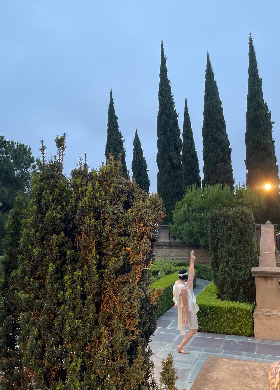 A dancer dressed as a flapper throws her leg in the air on the tile of a grand garden walkway. Tall cypress trees in the background