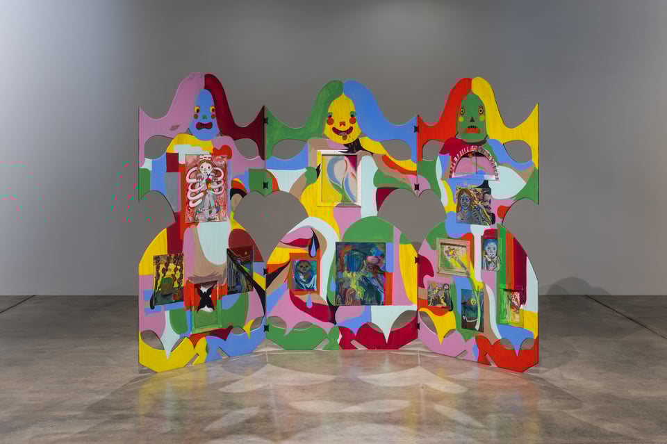 a wooden sculpture of three connected child figures, painted in bright colors and containing paintings on canvas, stand in a dim gallery