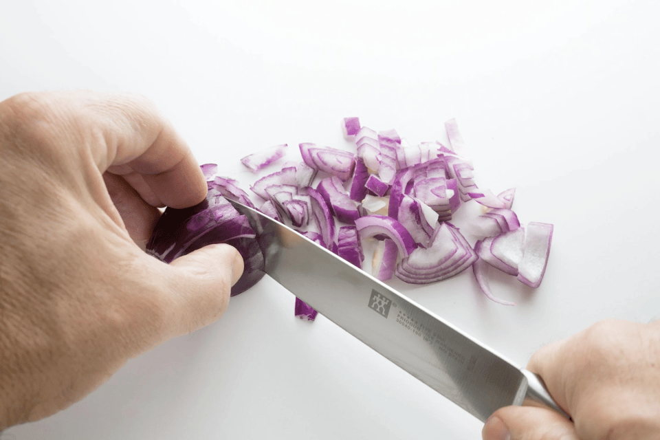 A close-up photo of a white man's hands chopping a red onion, on a white plain background.