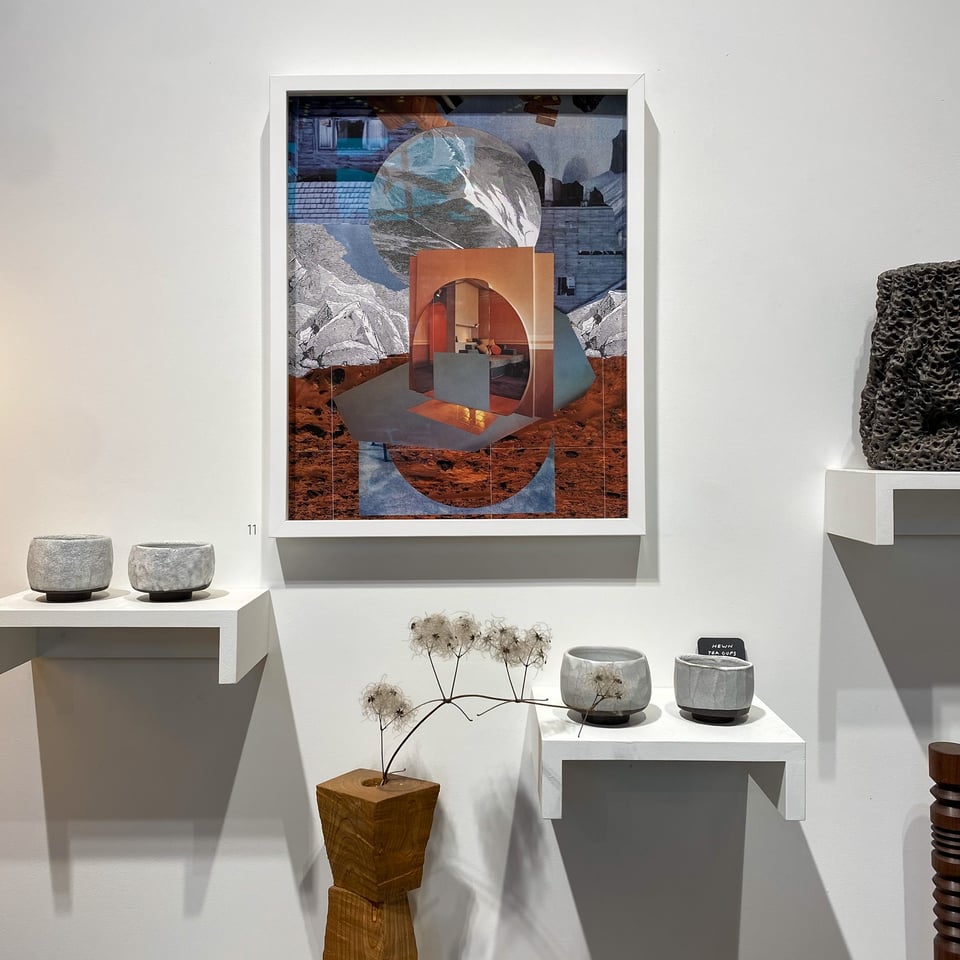 Installation view of Aimée Henny Brown's Thresholds at Propellor Studio's Granville Island gallery. A large framed collage work hangs centrally, combining geological landscape imagery with domestic interior spaces rendered in warm terracotta and cool grey tones. Stone ceramics on white wall-mounted shelves flank the piece; dried seedheads in a wooden block vase sit on a lower shelf below.