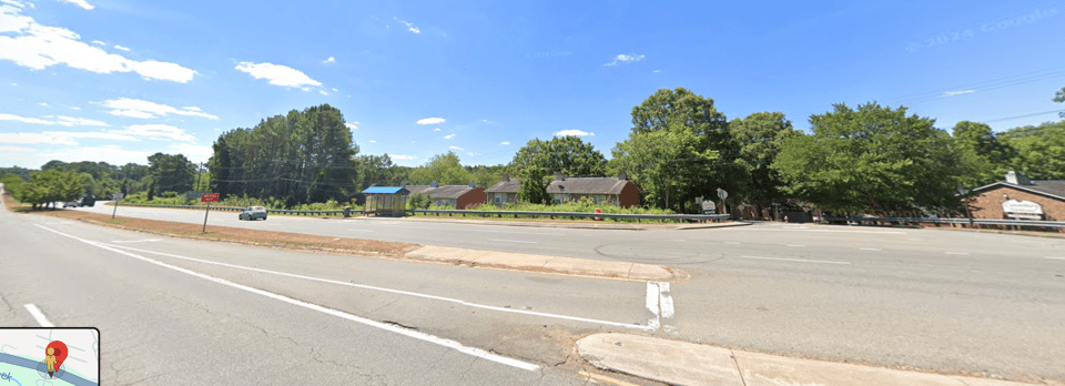 Cropped screenshot from Google Maps Street View on a bright partly cloudy day. The camera is in a Westbound lane of NC Highway 54 looking South at the intersection of 54 and Abbey Ln. A car traveling East is visible, as well as the roofs of several buildings behind a bus shelter near the center of the frame.