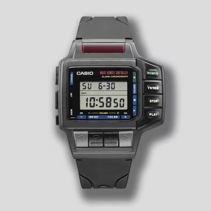 A vintage Casio wrist remote control watch is shown with a rectangular digital display and an array of buttons along the sides and bottom. Labels like “POWER,” “TV/VCR,” “STOP,” and “PLAY” indicate its ability to operate electronics, while directional and channel buttons sit below the screen. The black strap and chunky, gadget-filled design emphasize its 1980s–1990s tech aesthetic