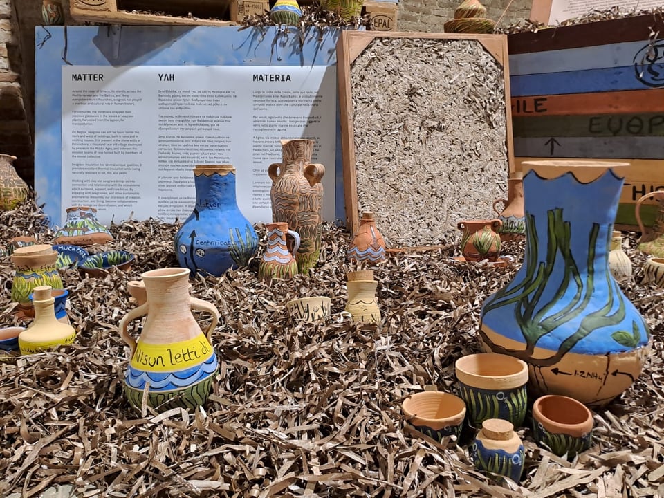 An image of Vessel's installation at the Venice Biennale: four brightly-coloured wooden crates are surrounded by piles of dried seagrass, in which are embedded over a hundred clay vases and other vessels, all painted with seagrass imagery and messages.