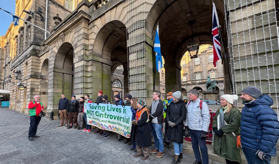 Sixteen adult protestors and two Edinburgh councillors - of varying ages and genders, mostly sporting warm winter clothes on a bright but chilly morning - stand outside the courtyard fascia of Edinburgh's City Chambers on the Royal Mile. Unlit christmas lights hang behind them, a saltire and union flag droop from their respective stantions, and the protestors hold up a wide canvas sign featuring Keith-Haring-esque abstract cycling figures reading 'Saving Lives is NOT controversial - LTNs, Modal Filters and Bus Gates NOW!