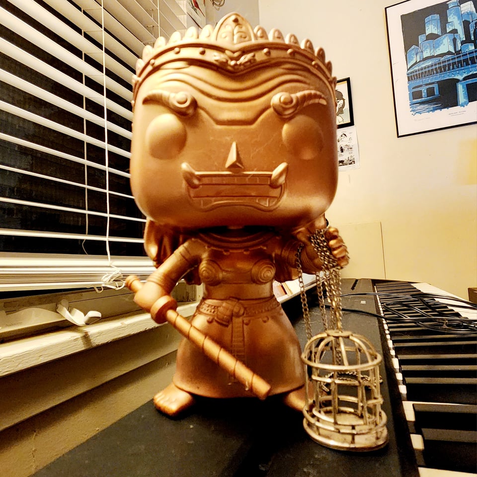A golden Funko POP! vinyl figurine, depicting the caricature of a Thai ogress, holding a baton in one hand.