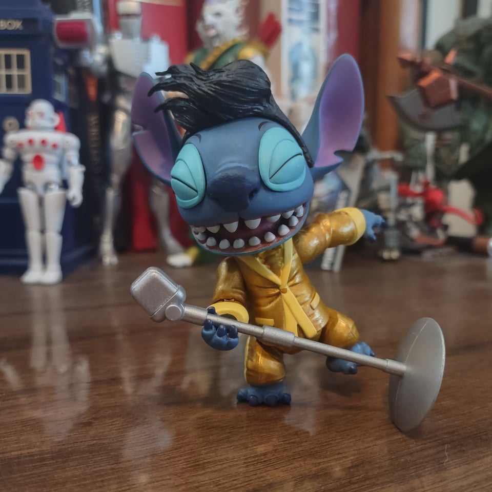 An action figure of the Disney character Stitch stands on a wooden desk with other action figures blurred in the background behind him. Stitch wears a gold lame tuxedo and somehow has a big, waving pompadour of black hair. he's balancing on one leg while he dips a microphone stand and sings into it.
