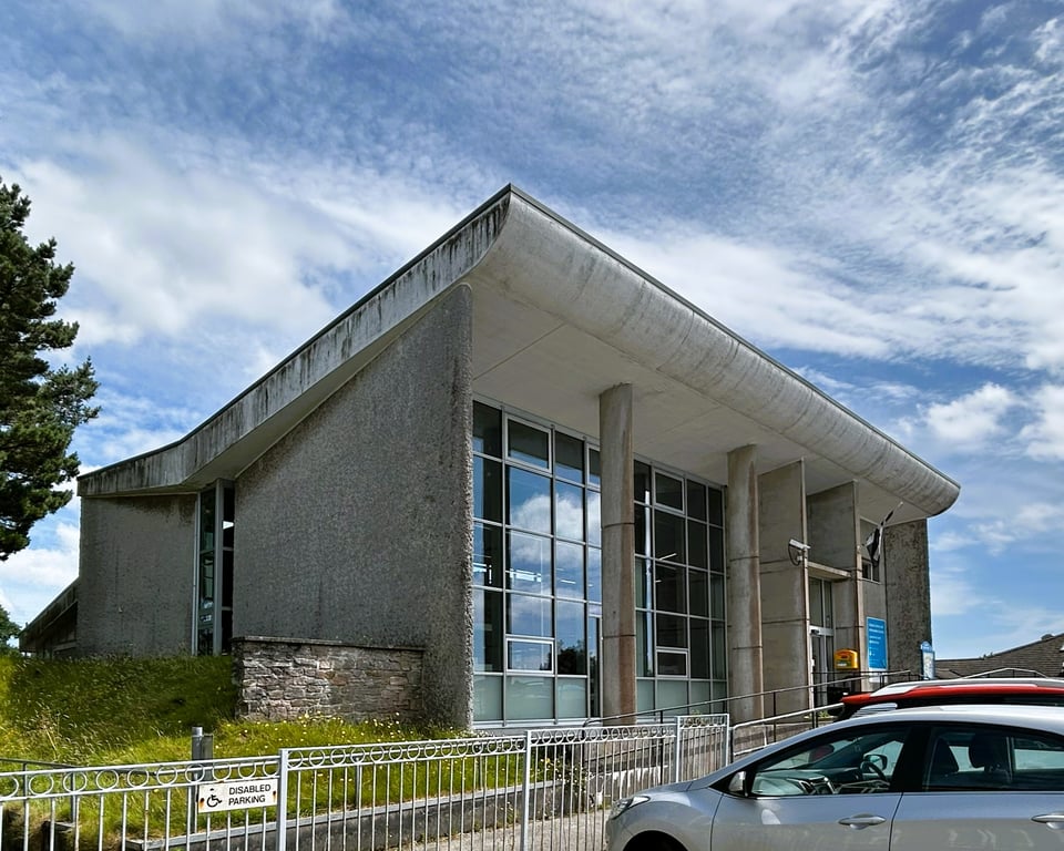 Concrete walls soar up to a double height butterfly roof. A curtain wall of glass is deeply recessed under the white swoop of the roof, and narrow oval pilotis hold it up.