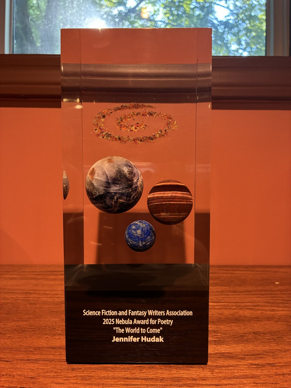 A clear rectangular obelisk with three gemstone planets and a sparkly swirled "nebula" embedded inside. The bottom of the obelisk is black, with gold writing that says, 'Science Fiction and Fantasy Writers Association / 2025 Nebula Award for Poetry / "The World to Come" / Jennifer Hudak'