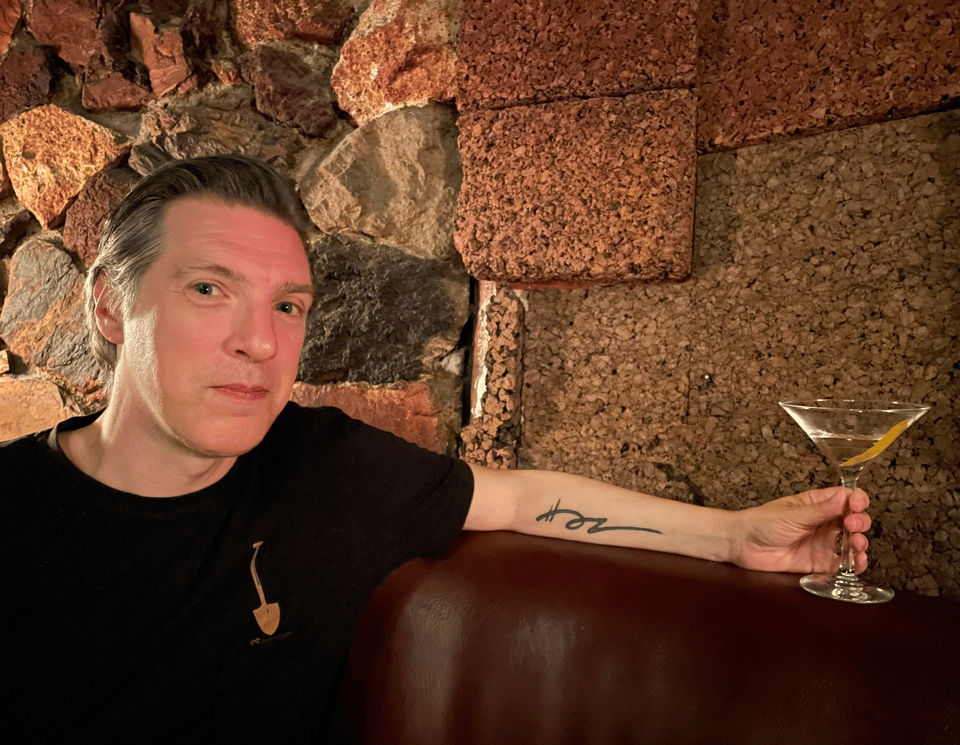A person smiling in front of a rocky wall, holding a cocktail and displaying a tattoo of a dancing line on his forearm