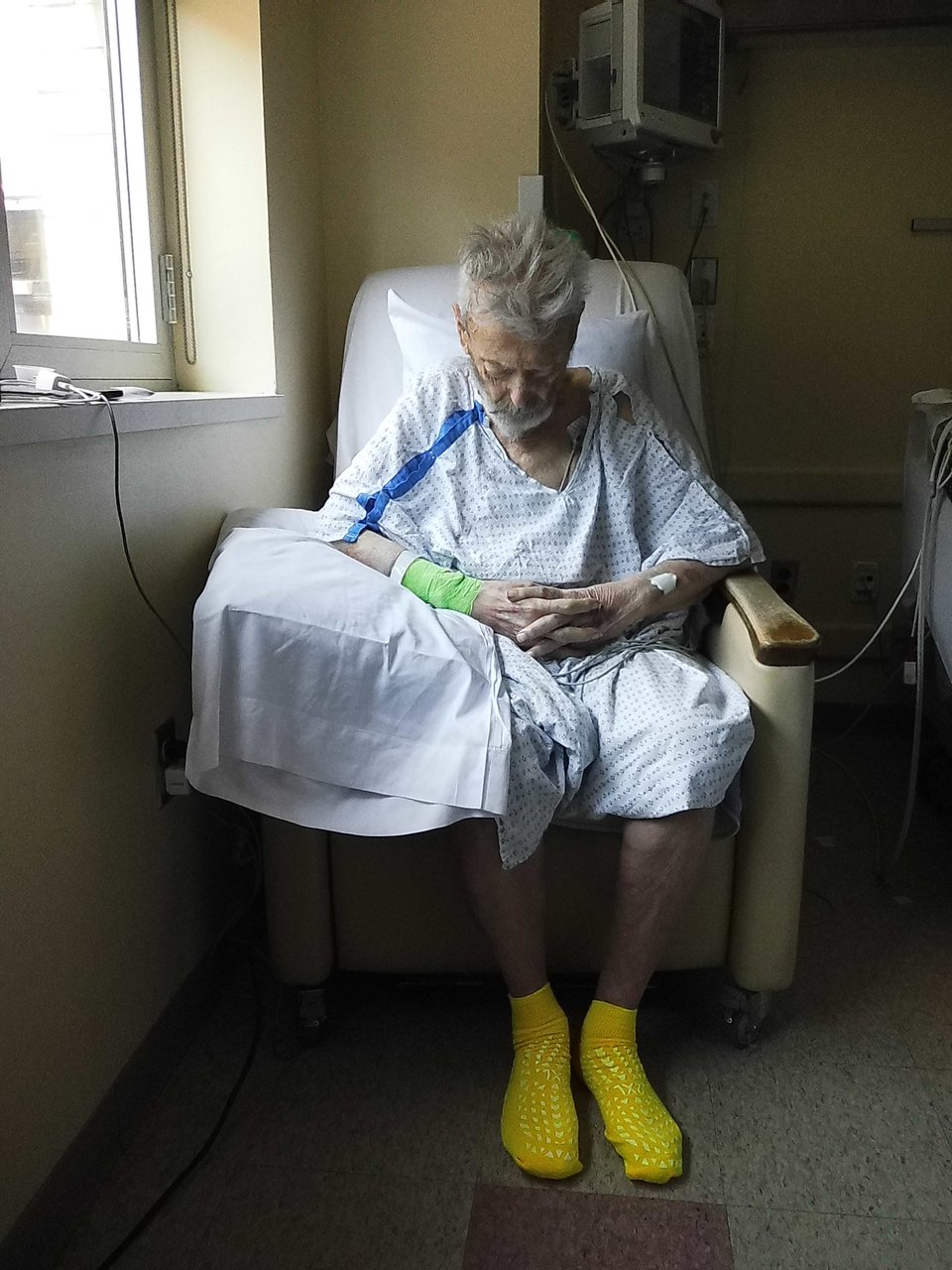 a digital Instax photo of an old white man with a beard sitting in a chair in a hospital room. His head is down and he's wearing a gown, with yellow safety socks on, and a pillow under his right elbow. The light is coming in from a window to his right, and there's a green wrap around his right wrist, keeping an IV line in place