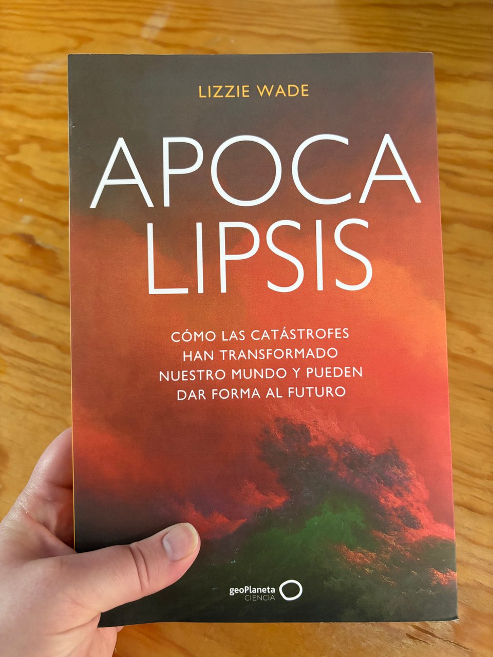 A hand holding the book APOCALIPSIS: Cómo las Catástrofes Han Transformado Nuestro Mundo y Pueden Dar Forma al Futuro. The cover is an image of orange and green clouds.