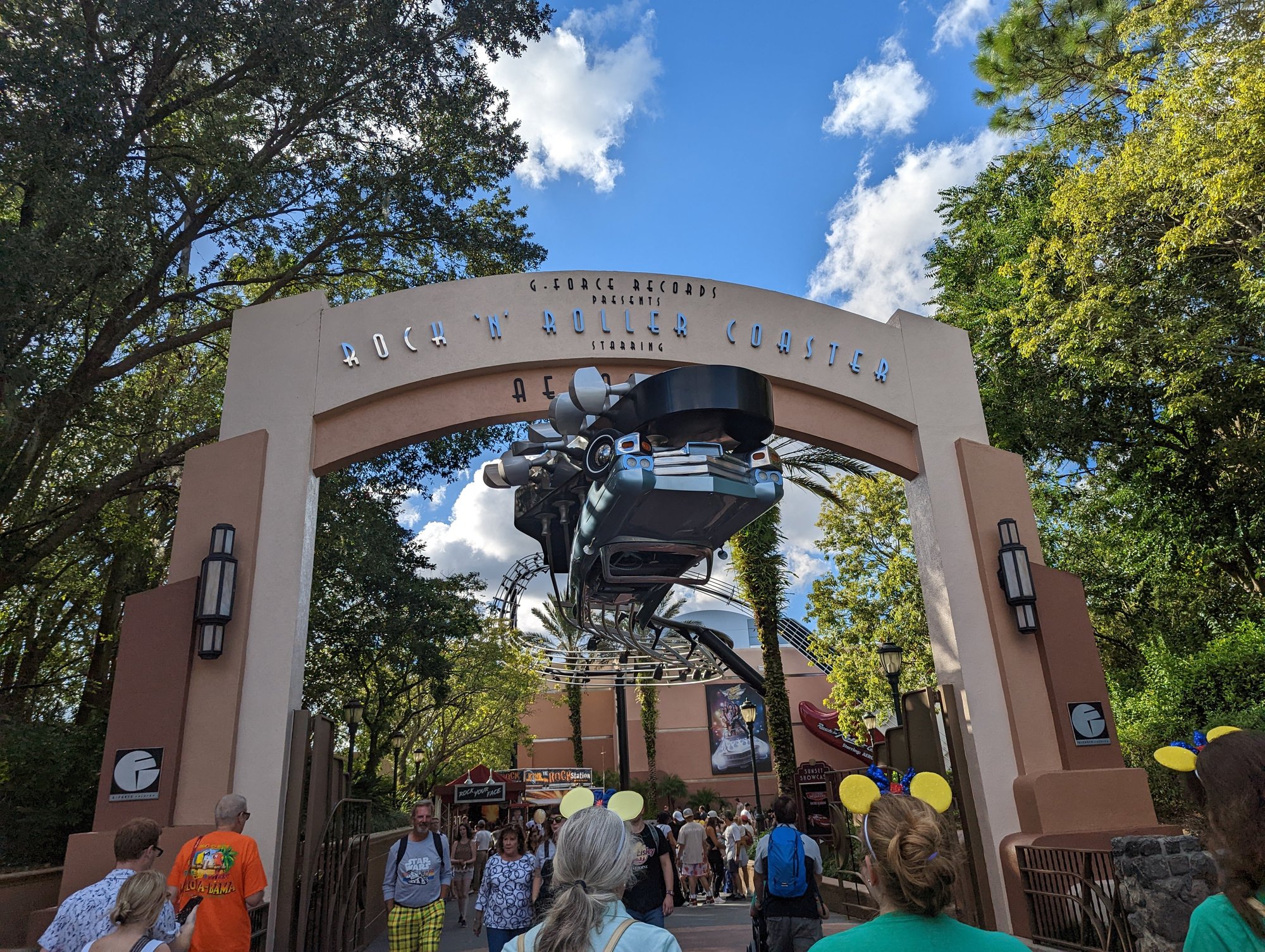 The entrance to the Rock n Roller coaster, with a powder blue limousine hanging on a track upside-down, and the track is also the headstock of a guitar with guitar strings floating through the air behind it. There are lots of people coming to ride the ride in the sunny afternoon