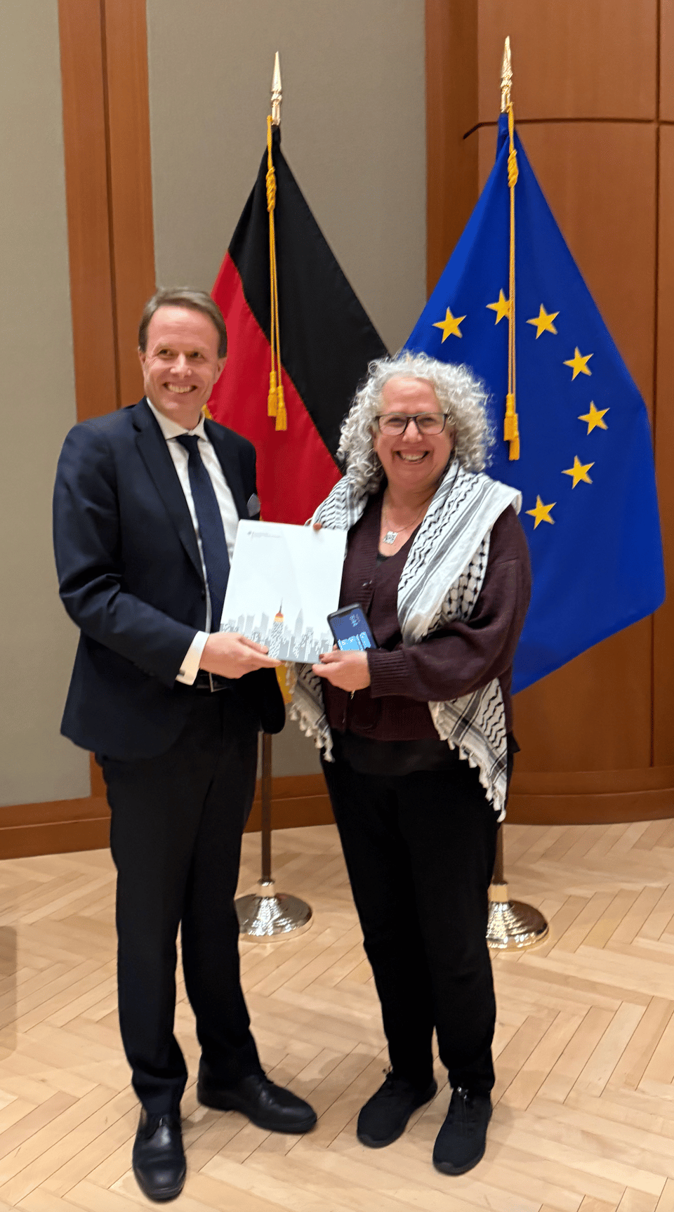 Photograph of a man and a woman, both smiling, standing indoors in front of German and European Union flags, holding a document together. The man is dressed in a dark suit. The woman has gray curly hair, glasses, a dark sweater and black pants,  and a black and white keffiyeh around her shoulders.