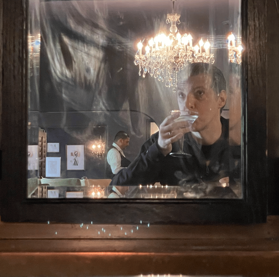 A man sits at a bar sipping a martini reflected in an old, foggy mirror