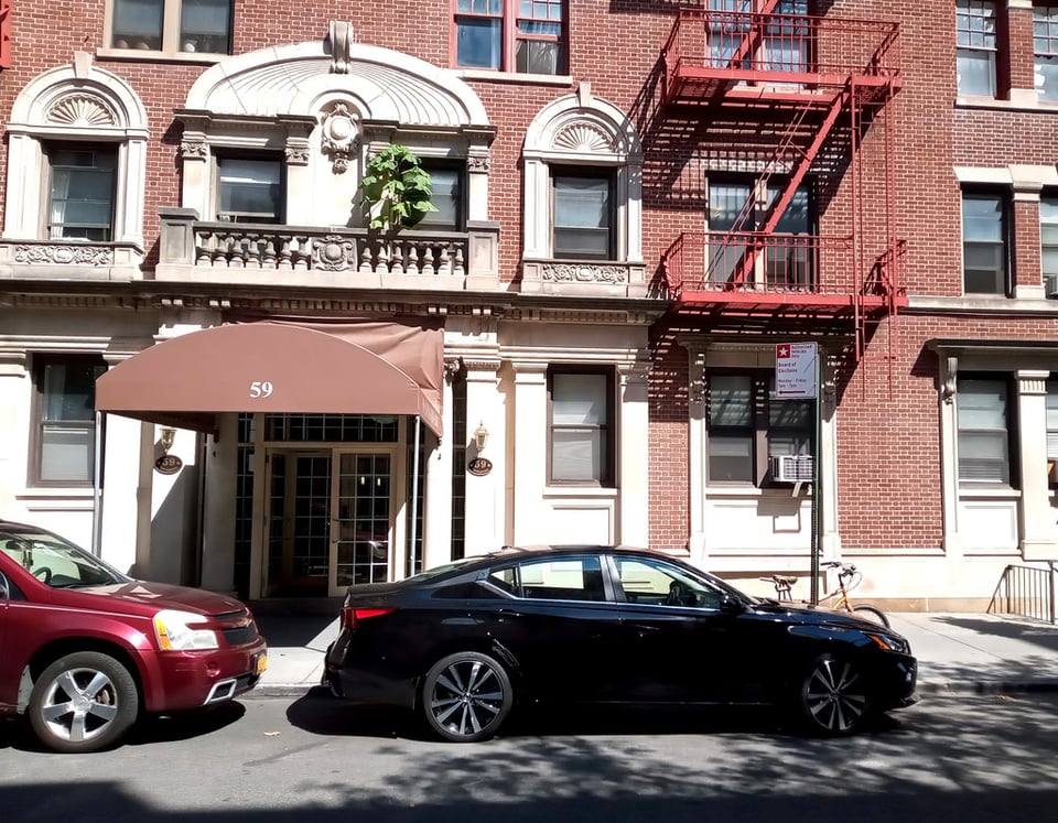 A photo of a brown and white residential building from across the street. There is a small green plant growing on a balcony in front of two windows over an awning..