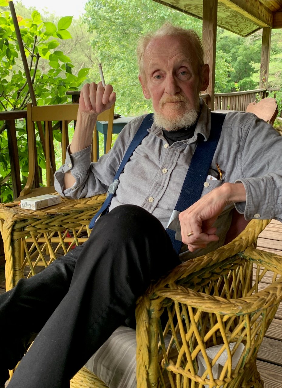 Photo of the late art critic Peter Schjeldahl sitting in a wicker chair on a deck, holding a cigarette in his right hand