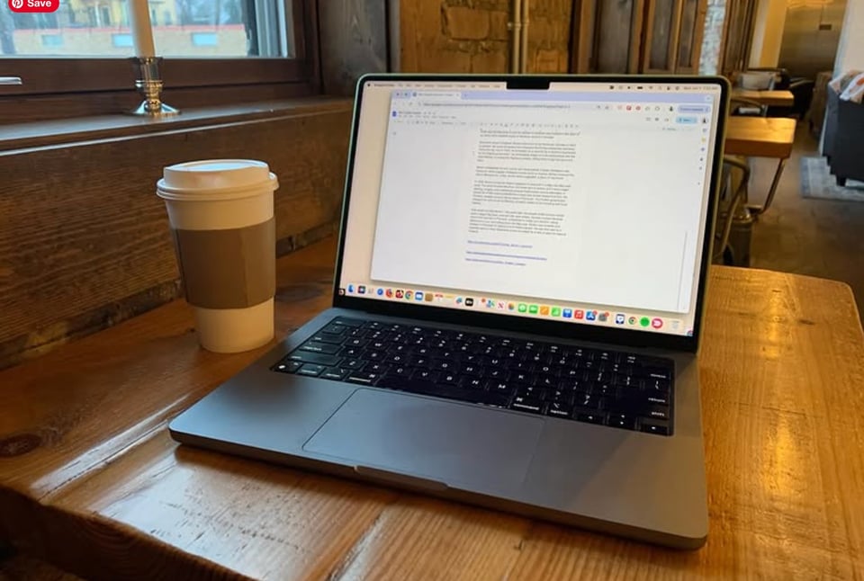 Image of an open laptop with an open word processing document on it. To the left of the laptop is a cup of coffee. The picture was taken inside a cafe.