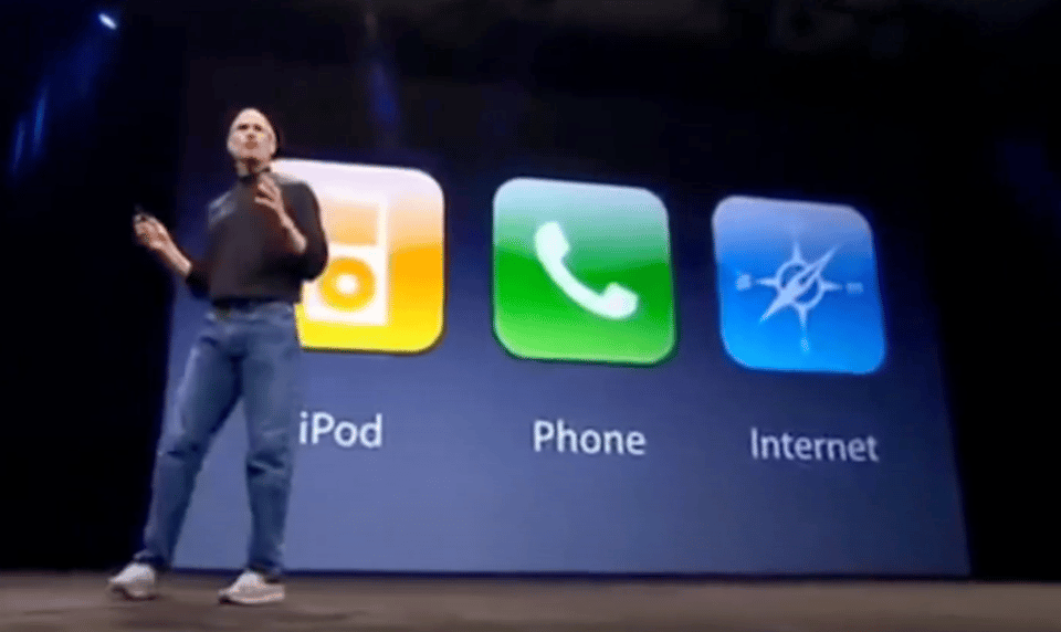 Steve Jobs in a black polo neck standing in front of a screen that says "iPod, Phone, Internet"