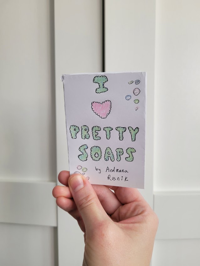 a light-skinned hand holding a mini paper zine titled "I heart pretty soaps" by Andreana Rosnik. The text on the cover resembles puffy bubbles, and the "I", "pretty", and "soaps" are blue-green. The heart is pink. There are pink, green, and blue bubbles on the border.