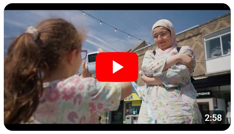Youtube thumbnail for a video about community events in Cambridge, showing a mother and daughter playing with a camera on the city street.