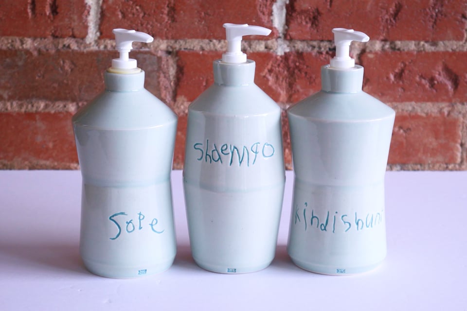An interlocking set of three angular soap dispenser bottles. The outer two angle in and back out, forming an hour-glass shape before coming in again to the bottle neck. The middle one angles out and back in, matching the angle of the others. They're glazed in a light blue, with darker blue inlaid child's handwriting that says sope, shaempo, and kindishinr
