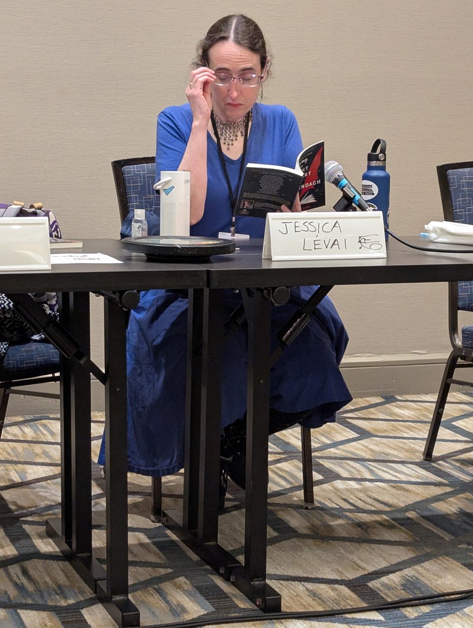 The author, in a blue dress, adjusts her glasses whle reading from her book, sitting at a table in a conference room.