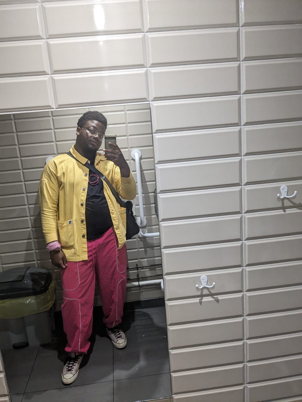 Tayo in a cinema bathroom mirror selfie wearing pink converses, pink tracksuit bottoms, a black t shirt, a yellow shacket and a pink bandana on left wrist.