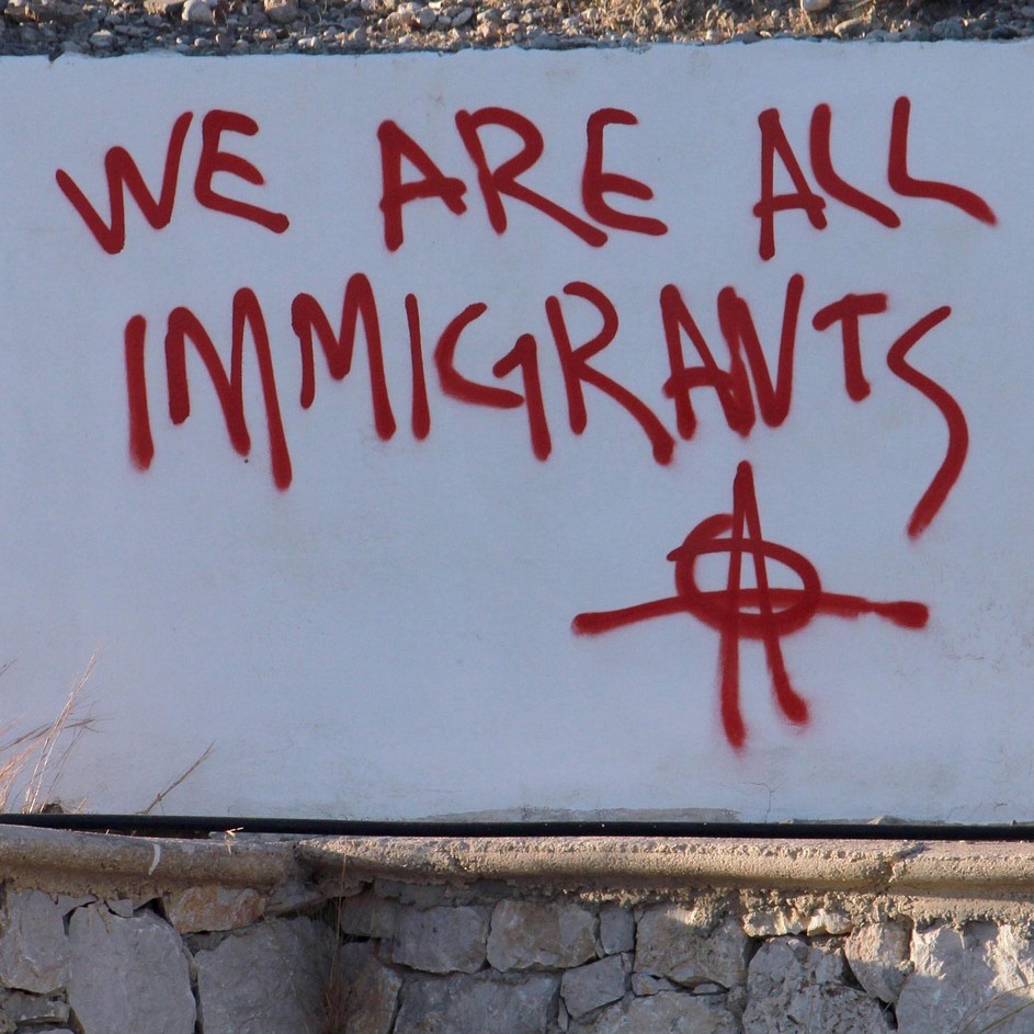 Red graffitti on a white wall. Text reads "We are all immigrants."
