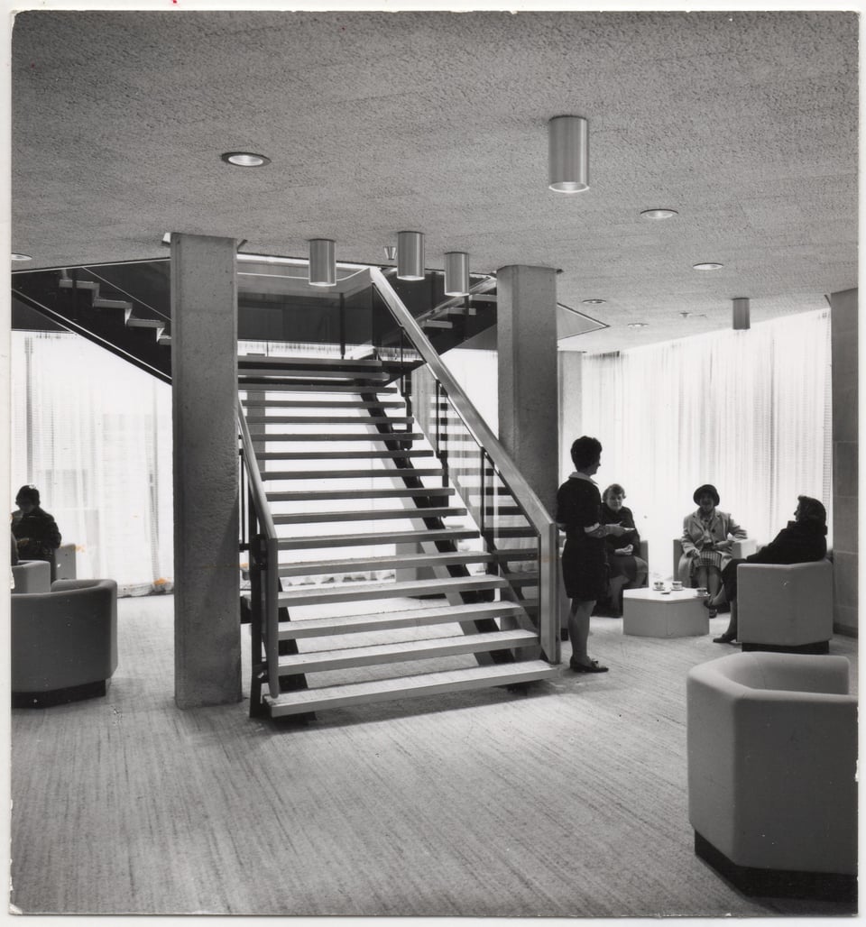 Interior with a large open staircase going up through an octagonal stair well. Clusters of octagonal chairs are organised in the lounge area, with octagonal low tables. Several women are having drinks whilst a server waits by the stairs.