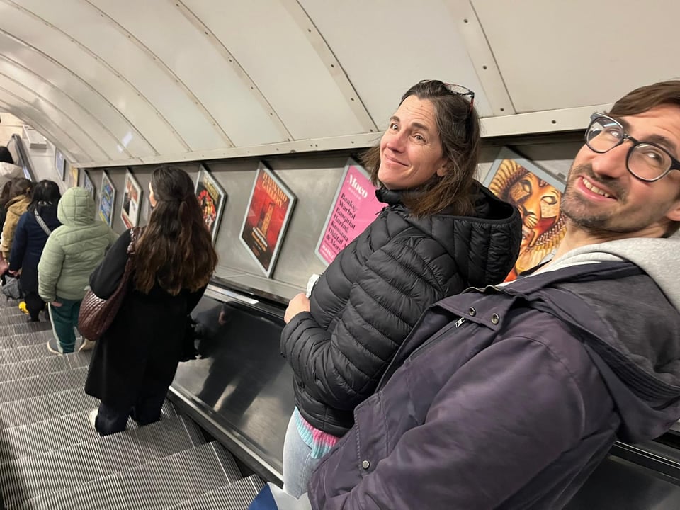 Two people going down the London Underground escalator smile at the camera.