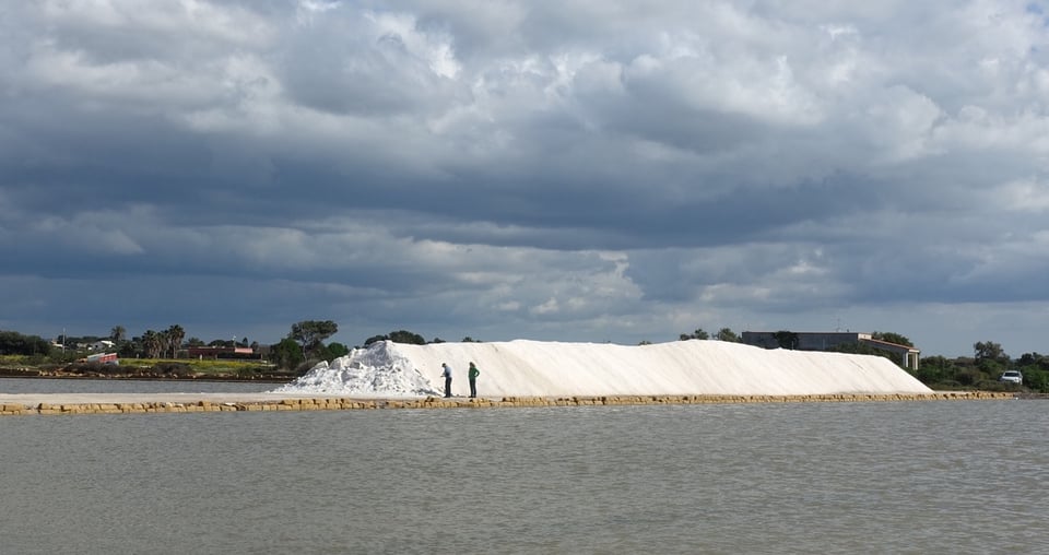 A long hill of salt, glistening white under a cloudy gray sky, dwarfs two workers in front of it. The foreground shows the shallow lagoon in Trapani, Sicily, where the water evaporates to concentre the brine.