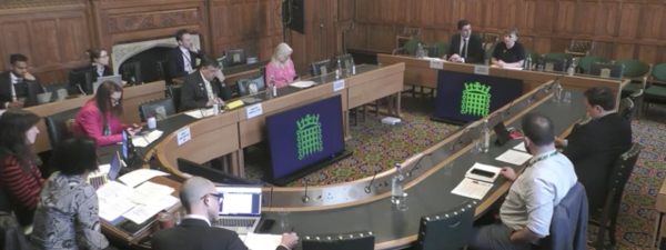 A parlimentary room with a large shoehorse table in the middle. 13 people are sitting around it with laptops and notes in front of them. The room has wooden walls and a patterned carpet. The civil service logo is presented on a screen in front of the tables.