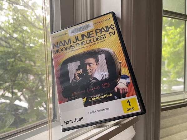 The plastic DVD case for “Nam June Paik: Moon is the Oldest TV” perched at an angle on a half-open window shade, with a view of green foliage beyond