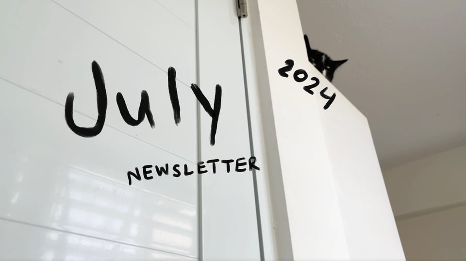 Cover for July’s video newsletter. There is a closet door to the left and a cabinet to the right. Mabel, a black and white cat, is peeking out from her bed on the top of the cabinet.