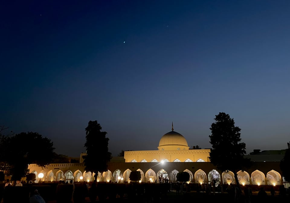 An almost panoramic picture of a mosque at night. Illuminated with many yellowish- warm lights. In the forground is a park/garden outside the mosque with people, and two tall trees prominent. In the background is a greyish/navy blue clear sky with a single bright star.