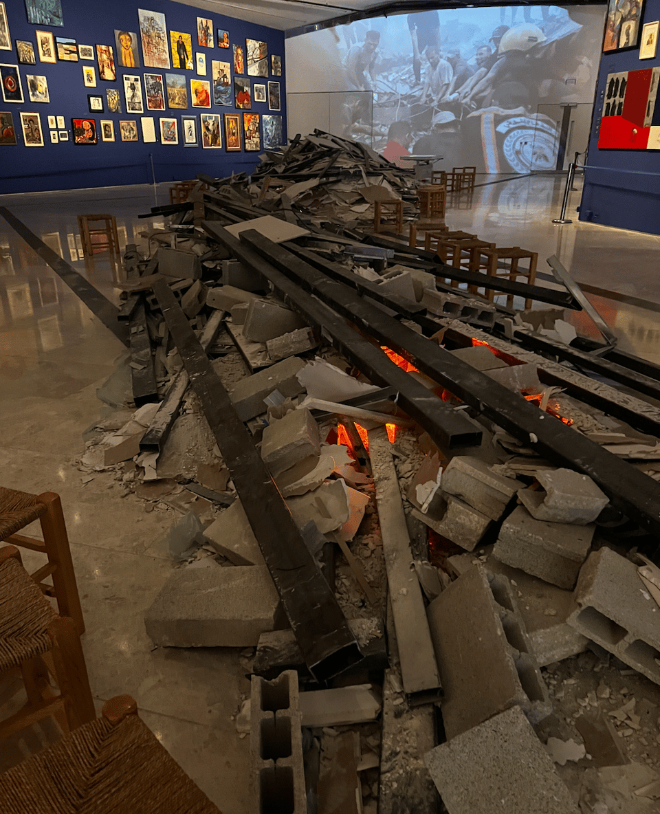 A blue wall with many paintings, rubble in the middle of the room, and on the far wall an image of rescue workers working in a disaster area