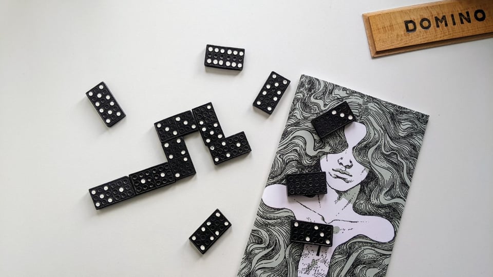 On a white surface lies a zine with a hand drawn cover of a figure obscured by a waterfall of flowing hair. They have a keyhole in their chest. There are also dominos there that form a short chain and dominos just scattered around, partly covering the zine.