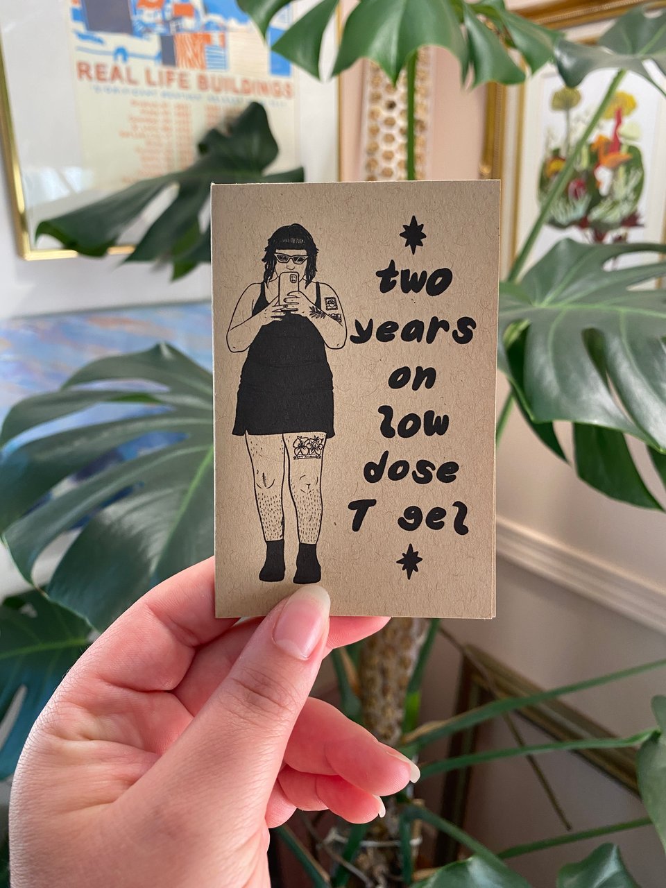 elliott holding up two years on low dose T gel mini zine in front of a big monstera plant.