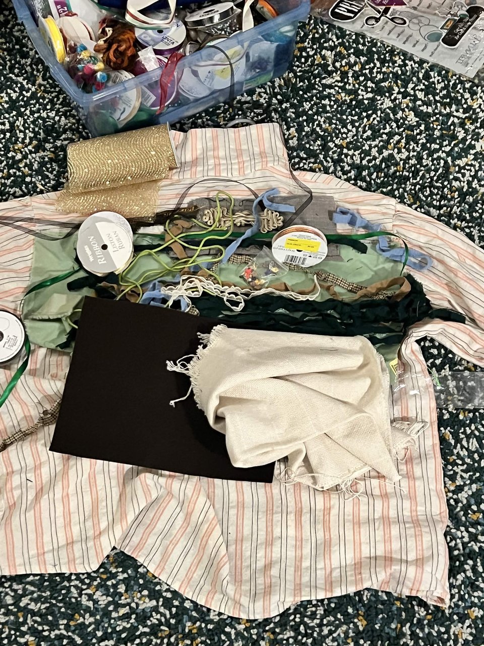 A set of fabric odds and ends placed on top of a pink grey and white striped shirt. Green fabric, brown felt, white lace trim, monk cloth, golden ribbon, green ribbon, grey tweed.