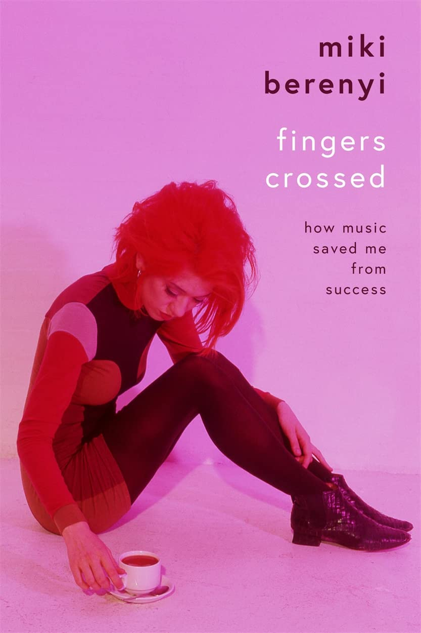 A woman with red hair and a black and red all in one long sleeve top and dress, reaches for a cup of coffee, head down, deep in thought.