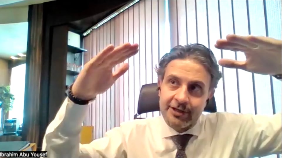 A man in shirt and tie before a window with blinds closed, his hands raised almost equally high at eye level.