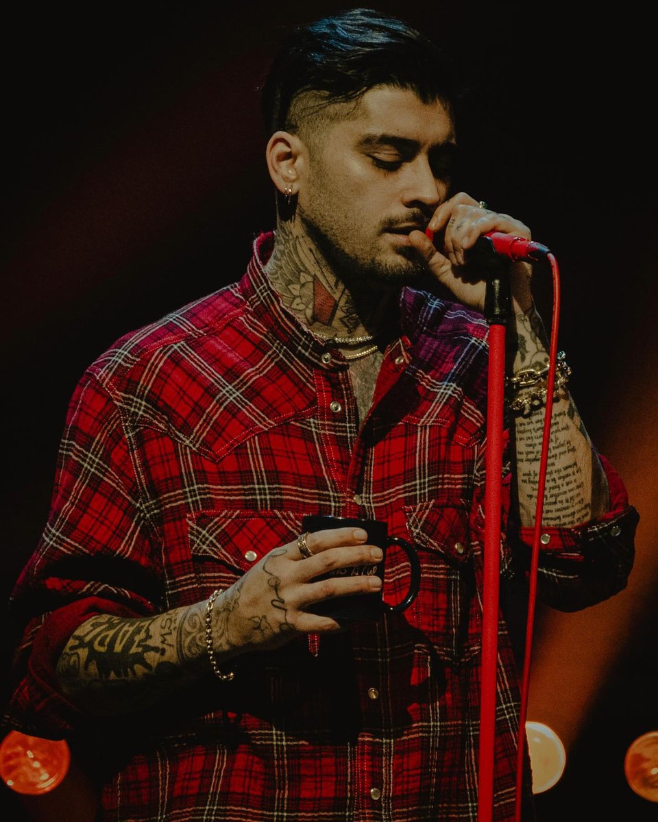 A handsome dark-haired man covered in tattoos and wearing a red plaid shirt holds a coffee cup and sings into a red microphone