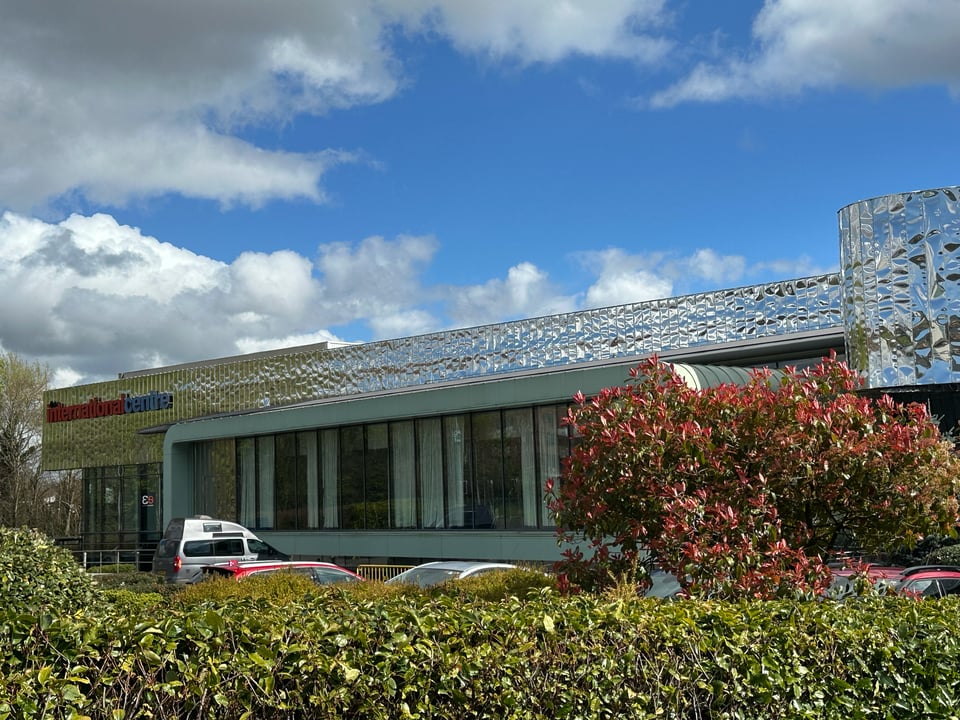 A picture of a long, lowish building, of a modern concrete and glass style. In front of it are lots of bushes and hedges. The upper part of the building is clad in a metallic covering that looks like slightly crumpled tin foil.