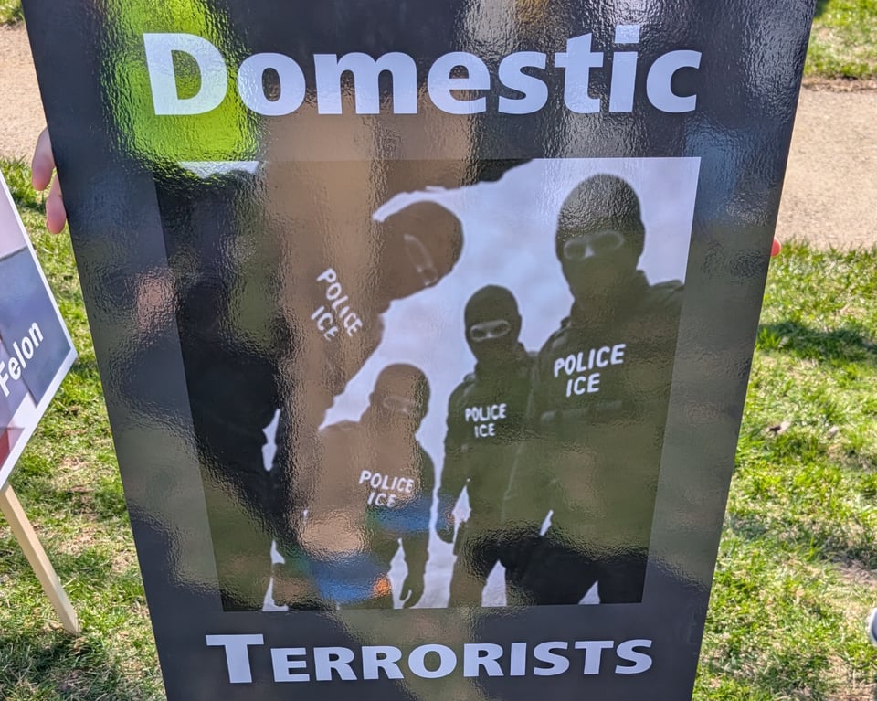 A sign with black background and white letters: A photo of militarized agents wearing masks with Police ICE on their vests with the words Domestic Terrorists above and below the photo.